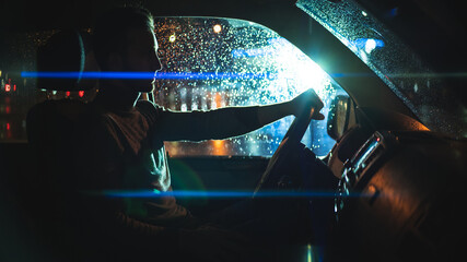 The handsome man is sitting in the car on the night rainy street