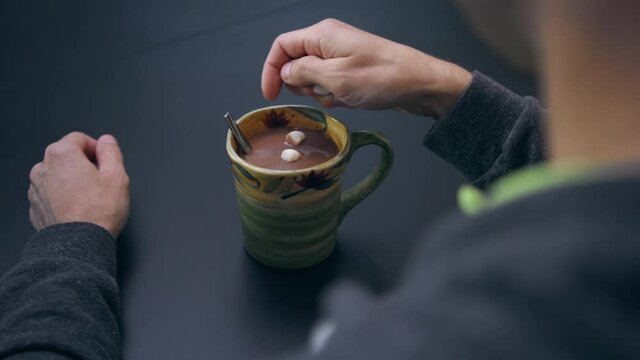 High quality close-up 4k slow motion video of a man mixing hot chocolate beverage in his cup with small white marshmallows view from the back. Handsome male drinking hot chocolate with marshmallows.