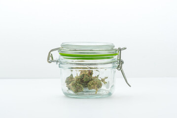 Marijuana in a glass jar with a green seal with lid closed.  Legal weed from a dispensary on a white surface, with white background, and subtle gradients.  Space for copy.