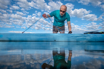 senior male stand up paddler is attaching a safety leash to his paddleboard before morning paddling workout, low angle view in blue tones from a partially submerged camera