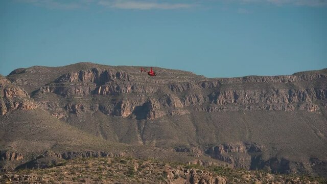 Red helicopter flying over mountainous landscape in the desert with clear blue sky in the background