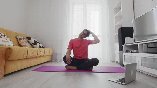 Young transgender homosexual man with make up stratching neck on mat at home with help of his instructor online on laptop at home