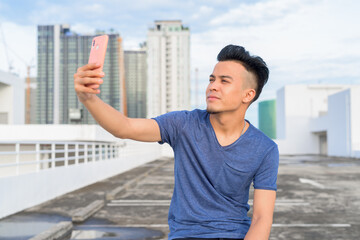 Young handsome multi ethnic man sitting and taking selfie against view of the city