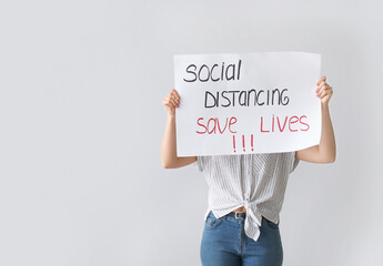 Young woman holding paper with text SOCIAL DISTANCE. SAVE LIVES on light background. Concept of epidemic
