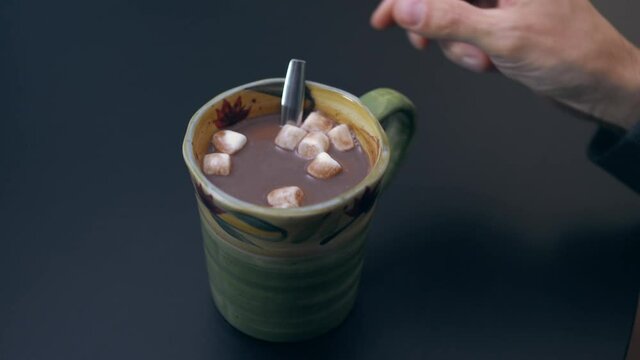 High quality close-up 4k slow motion video of a man mixing hot chocolate beverage in his cup with small white marshmallows view from the back. Handsome male drinking hot chocolate with marshmallows.