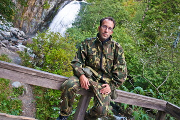 Portrait of a brutal tanned man, guy, brunette, with bristles and glasses in the wild, who sits in...