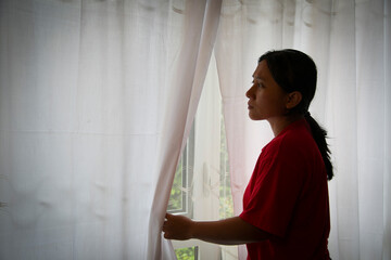 A woman anxiously peeks through her curtains in dark room. Suitable to illustrate fears stirred by...