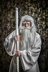 A grey-haired bearded severe wizard in a white cloak and a cap with a staff against a golden...