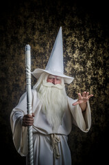 A grey-haired bearded severe wizard in a white cloak and a cap with a staff against a golden...