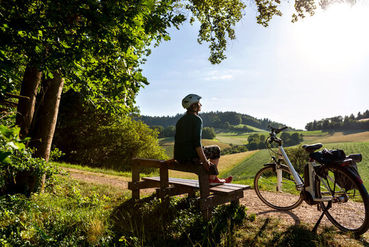 Frau genießt nach anstrengender Fahrradtour mit Ihrem Elektro Fahrrad die Abendsonne auf einer Holzbank in Idyllischer Hügellandschaft