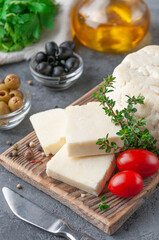 Cut Adyghe cheese into pieces. Cheese on a wooden board. Served with thyme, spices and cherry tomatoes. In the background are olives, parsley and olive oil. Background gray concrete.