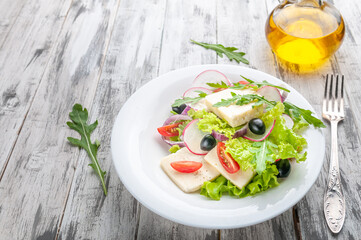 Salad with Adyghe cheese, arugula, olives, radishes, red onions and cherry tomatoes. On a white plate. Near a fork and, olive oil. Gray wooden background.