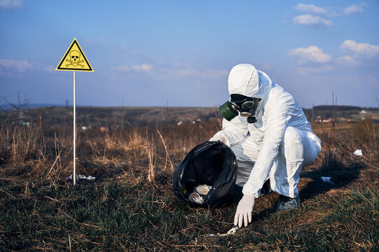 Male environmentalist in protective suit and gas mask putting trash into garbage bag with warning symbol of poisonous substances and danger. Concept of ecology, environmental pollution and hazard.