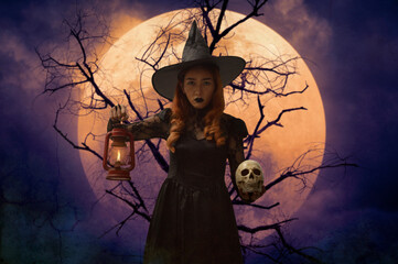 Halloween witch holding ancient lamp and skull standing over dead tree, full moon and spooky cloudy...