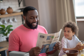 Dark-haired african american father reading a newspaper while having breakfast with his daughter