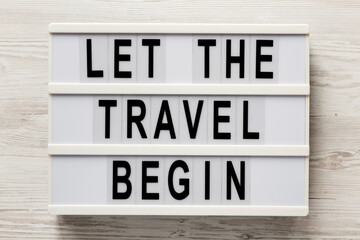 'Let the travel begin' on a lightbox on a white wooden background, top view. Flat lay, from above, overhead.