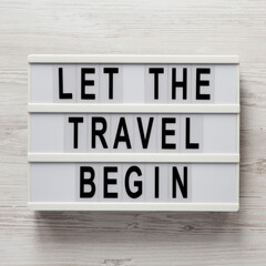 'Let the travel begin' on a lightbox on a white wooden background, overhead view. Flat lay, from above, top view.