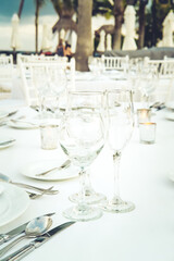 Festive serving with glasses for an outdoor celebration with garland lamps and palms at the background. Wedding, anniversary, birthday dinner and party. Toned image with place for text