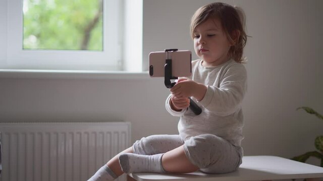 Toddler girl sitting at home and using selfie phone stick. Influencer, social media for kids