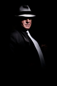 Man in a dark suit, a white tie and a white hat is emotionally posing against a black background in the image of an Italian mafioso in retro style.