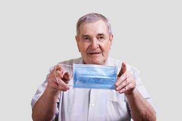 An elderly man on a gray background holds a blue medical mask in front of him. The concept of respiratory diseases, influenza, bacteria, viruses, pandemic, COVID-19.