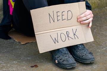 unemployed in old shoes holds in his hand a cardboard tablet with the inscription need work on the ground are small coins of alms.