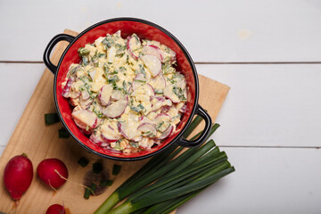 Radish, green onions, parsley, eggs salad in red bowl. Some fresh vegetables and greenery on kitchen chopping board on white wooden background. Top view, copy space.