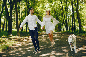 Obraz premium A beautiful and gentle girl with light hair and a white dress is walking in a sunny summer forest with her handsome guy in a white shirt and dark pants and they playing with cute gray dog