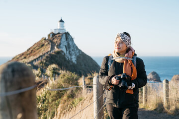 Beautiful asian tourist take some picture at Nugget Point, Dunedin, New Zealand. Young asian traveller enjoys walking in morning along coastline of New Zealand. Lifestyle image with natural landscape.