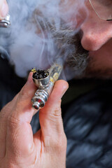 Male middle-aged with grey beard and mustache puffs a marijuana pipe with smoke in the background.
