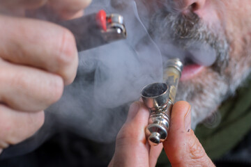 Horizontal view of ale middle-aged with grey beard and mustache puffing a marijuana pipe.