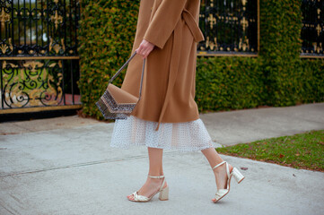 Detail of young fashionable woman wearing beige coat, tulle midi skirt and gold high heels. She holding stylish beige handbag with silver details in hands. Street style.