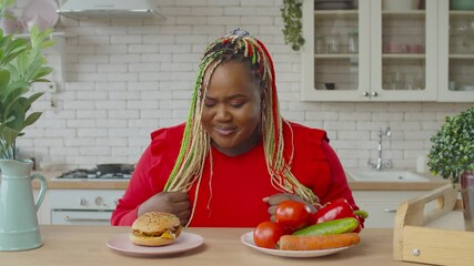 Confused charming overweight african woman with afro braids choosing between healthy and junk food in domestic kitchen. Black obese female on diet can't resist craving to eat tasty hamburger indoors.
