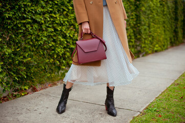 Detail of young fashionable woman wearing beige wool coat, tulle midi skirt and black high heel cowboy boots. She is holding stylish burgundy handbag in hands. Street style. - obrazy, fototapety, plakaty