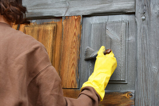 Teenage girl with brush painting old aged wooden garage doors with grey protective paint, back view.  DIY renovation concept.
