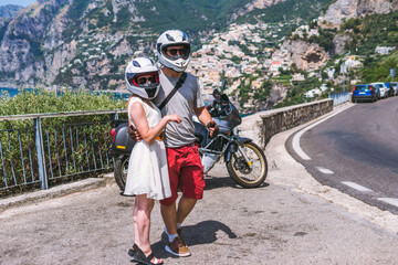 Fototapeta premium A couple of motorbike travelers poses against the backdrop of an incredibly beautiful view of Positano, a city on the edge of cliffs, the sea and a winding road. Travel and tourism. Active lifestyle.
