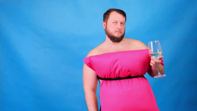 Funny bearded guy in a pink dress made of pillows drinks wine from a glass on a blue background. Crazy quarantine. Fashion 2020. Put on a pillow. Challenge 2020 due to house isolation
