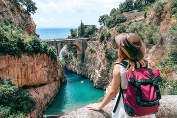 A girl in a cowboy hat with a backpack enjoys an incredibly beautiful view of a stone bridge over the gorge Fiordo di Furore. Little bay with paradise beach. Back view. Amalfi, Italy - obrazy, fototapety, plakaty