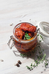 Homemade sun-dried tomatoes in oil with fresh herbs (thyme, oregano) in a glass jar on a light wooden background, soft selective focus
