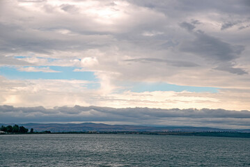Gray rain clouds hang low over the calm surface of the sea. Forest and hills in the distance.