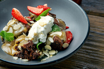 Appetizing brownie dessert cake with ice cream, strawberries and almonds in a gray bowl on a wooden background