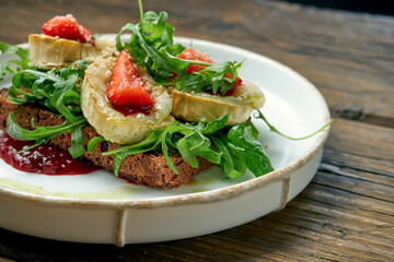 Appetizing toast with goat cheese, red jam, arugula and strawberries with rye bread on a plate on a wooden background