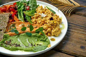 An appetizing vegetarian dish is scramble from tofu cheese, a salad of baked peppers and arugula and two types of hummus. Wood background