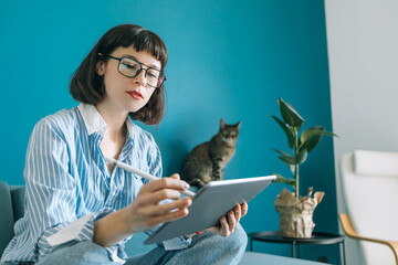 Beautiful brunette with a short haircut works in the home office on a blue background. Typing on a laptop, calling on the phone, transferring documents, making notes, drinking coffee from a mug.