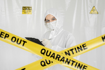 Epidemiologist woman in protective clothing is in a restricted area with a planchette. Yellow line Keep Out Quarantine. Quarantine alert sign. Entrance is forbidden in quarantine zone