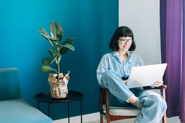 Beautiful brunette with a short haircut works in the home office on a blue background. Typing on a laptop, calling on the phone, transferring documents, making notes, drinking coffee from a mug.
