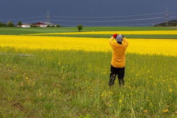 A man stands in a field of rape taking a photograph of the landscape on a showery day in Germany