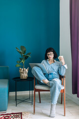 Beautiful brunette with a short haircut works in the home office on a blue background. Typing on a laptop, calling on the phone, transferring documents, making notes, drinking coffee from a mug.