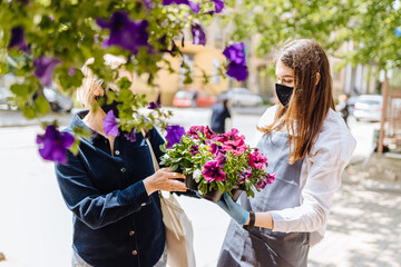 Young woman florist helping female client to choose potted plant in flower shop.Females owner of floral shop and consumer inprotective masks in faces.Lockdown, quarantine, back to normal concept