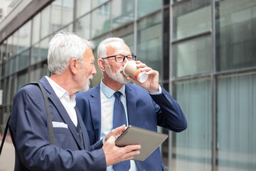 Two happy gray haired senior Caucasian businessmen walking down the street, drinking coffee and discussing business plans